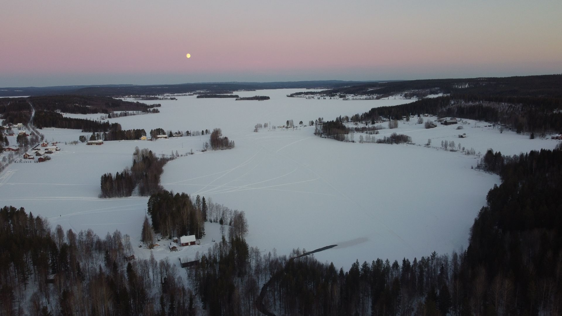 Aerial view of a frozen lake surrounded by snowy forest at dusk, with a faint moon in the sky