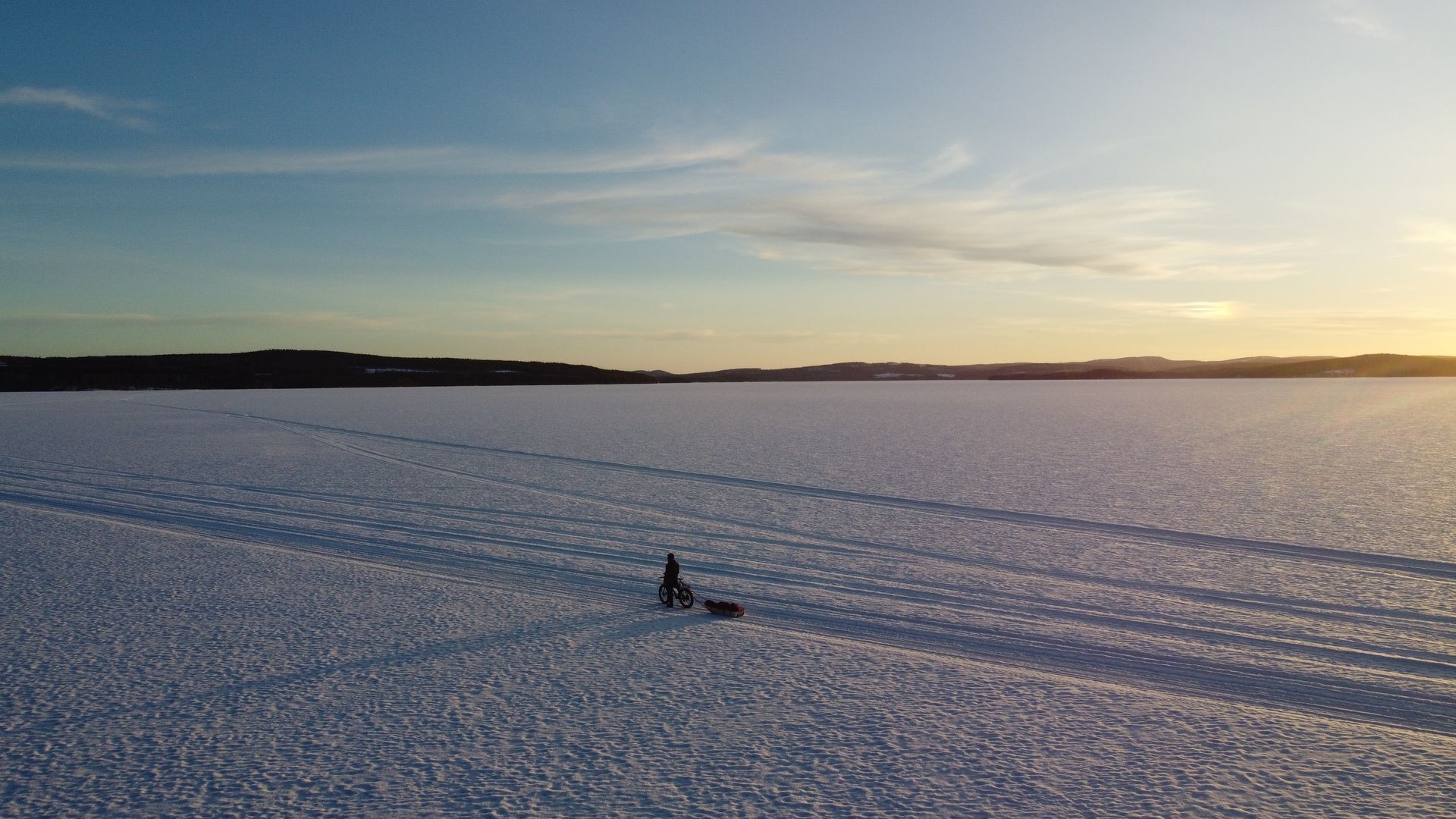 Wide snowy field at sunset with a lone person standing near a sled or snowmobile in the distance