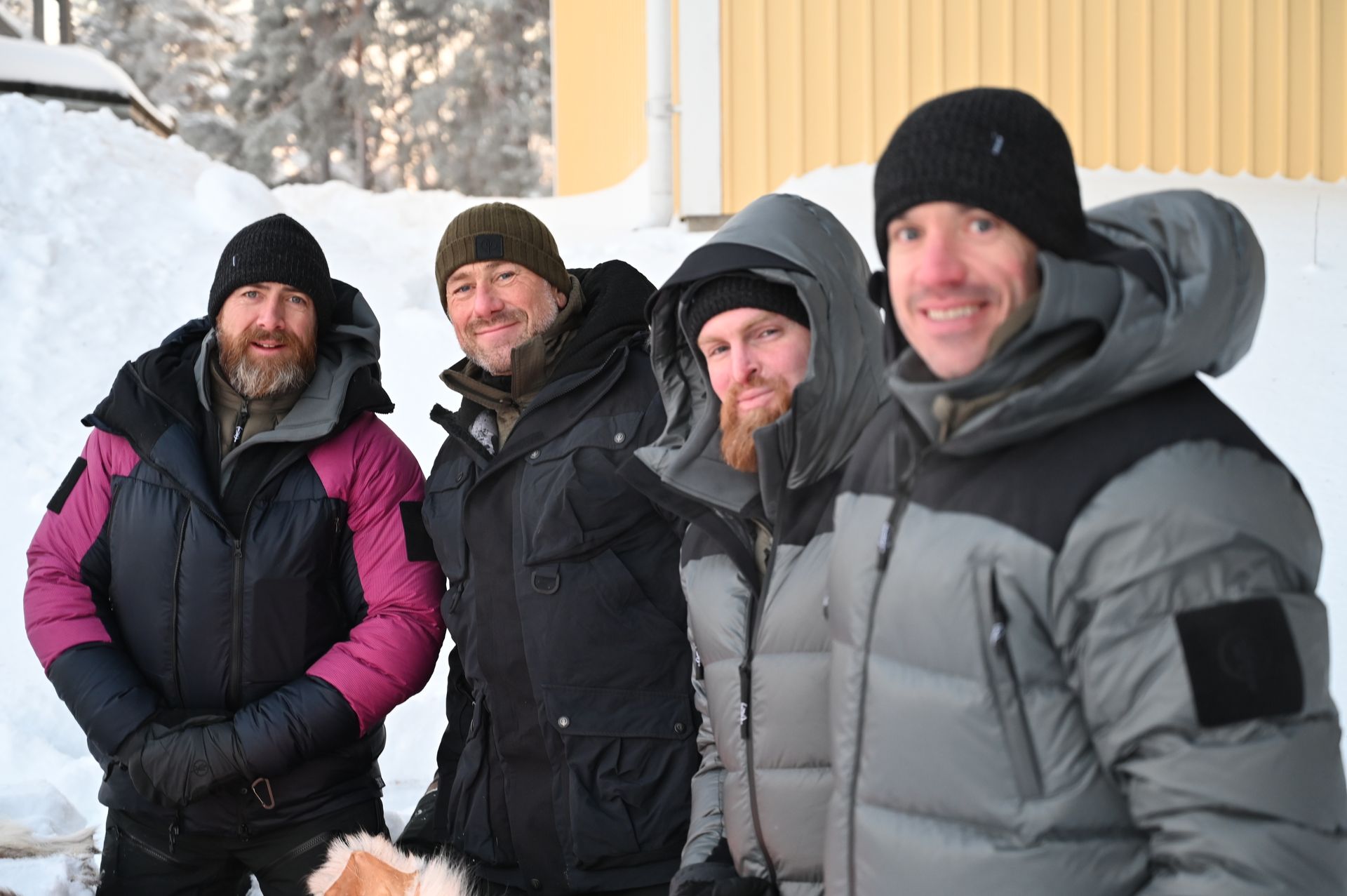 Four bundled-up hikers smiling in snowy woods, wearing winter jackets and beanies.