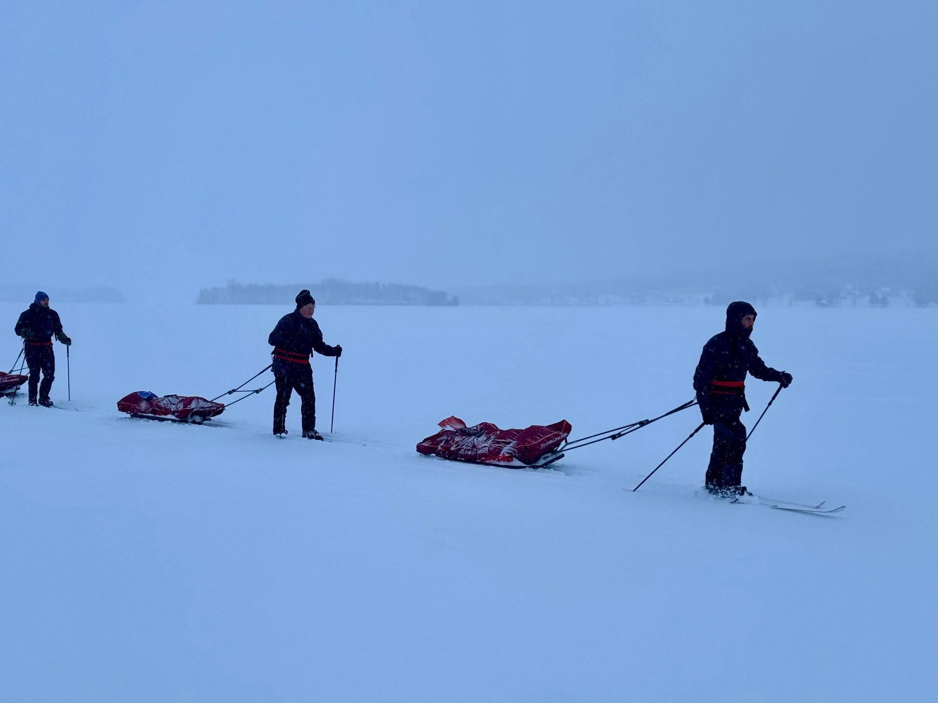 Three skiers towing sleds across a snowy, foggy landscape