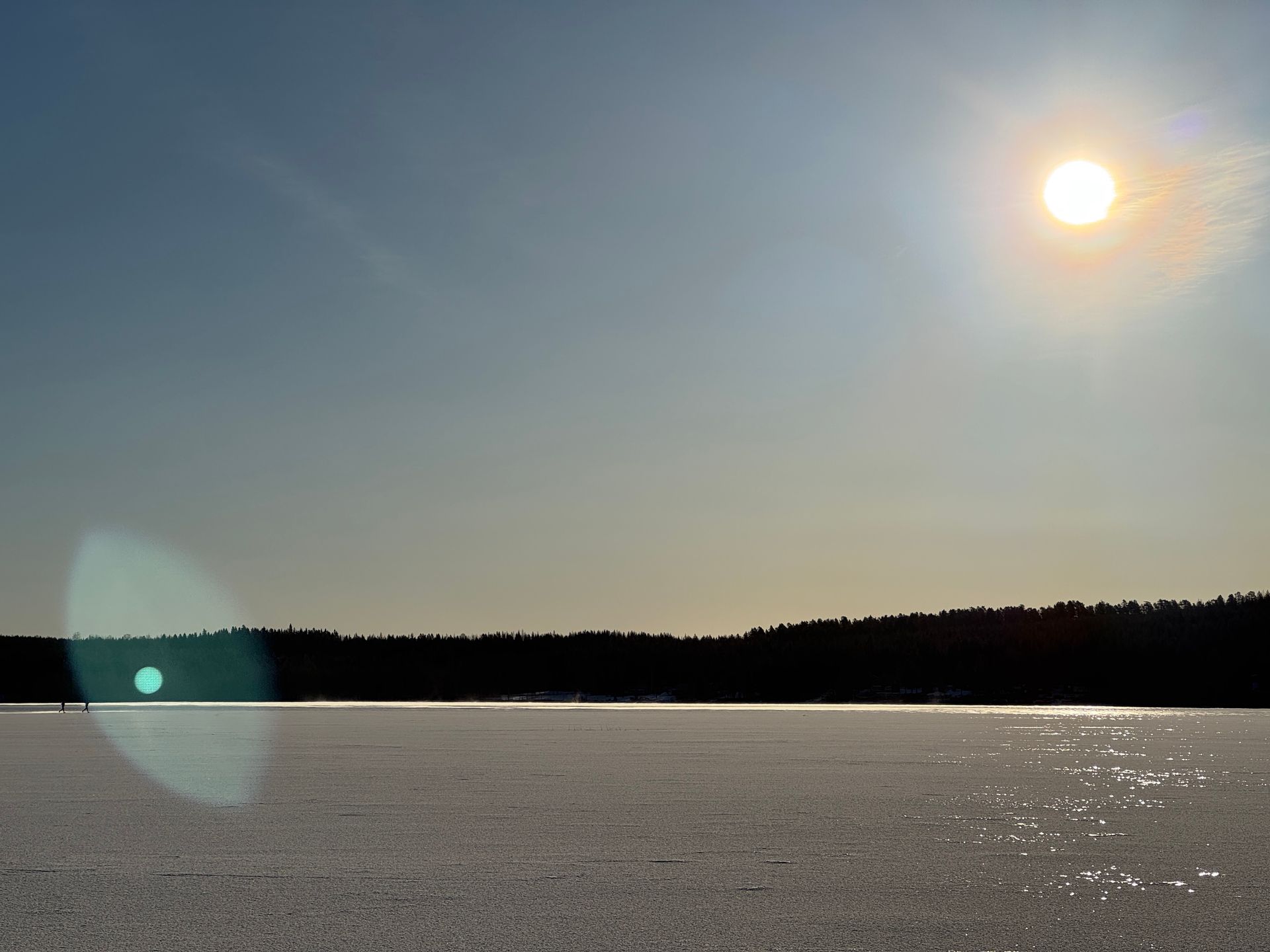 Sun shining over a calm frozen lake with a dark treeline on the horizon