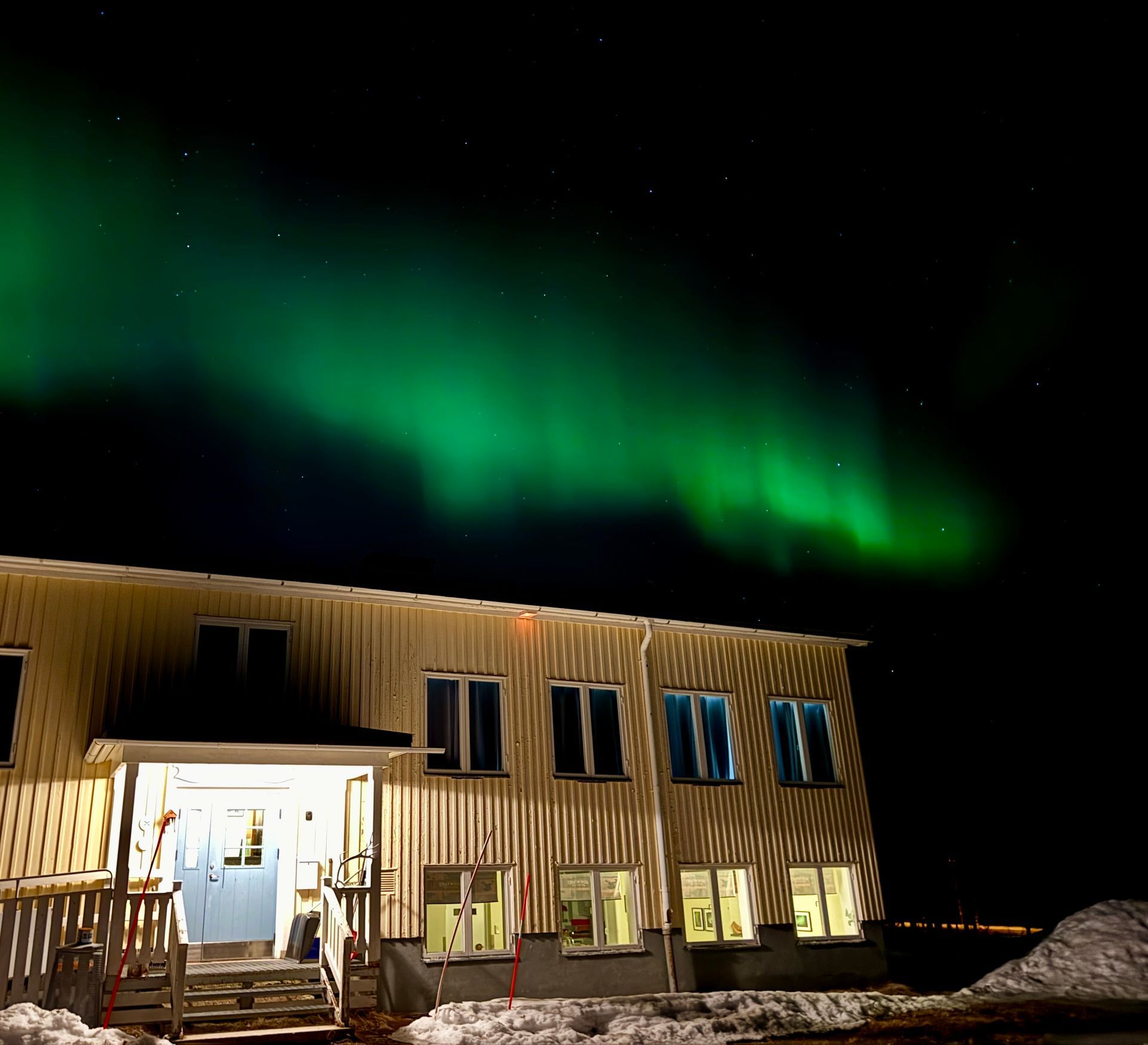 Green aurora over a lit wooden building at night in a snowy landscape