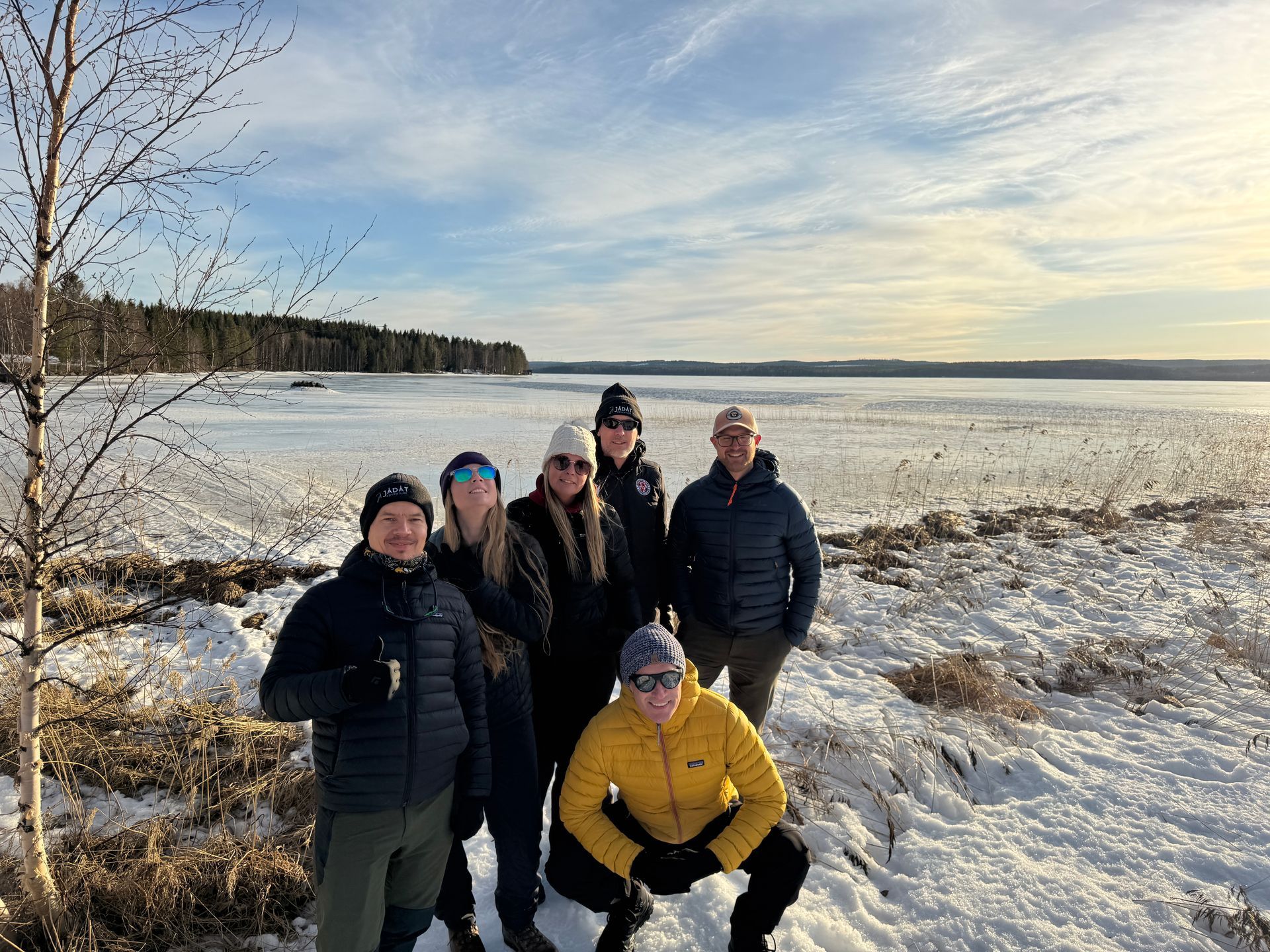 Six people posing on a snowy lakeshore under a cloudy sky, one in a bright yellow jacket.