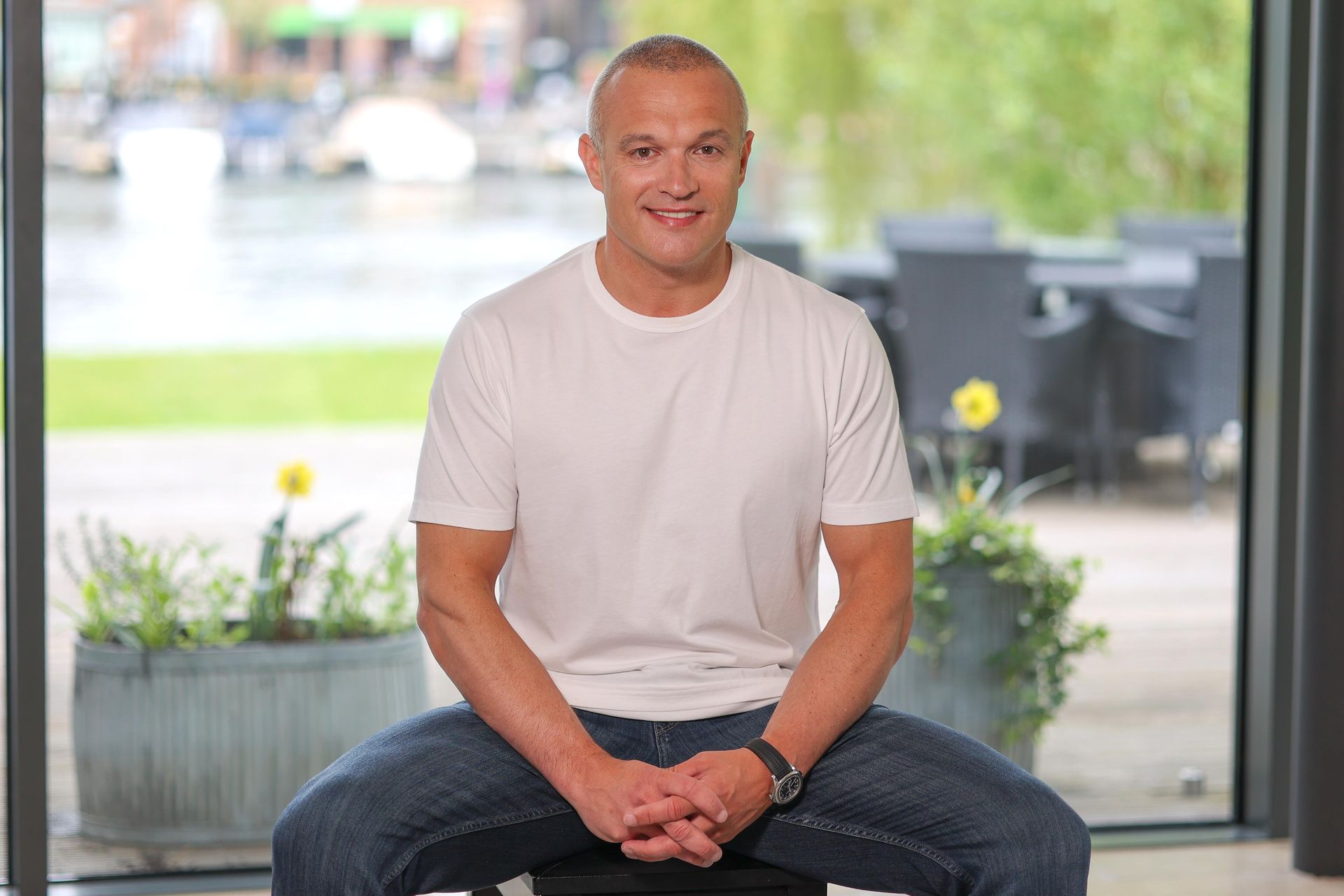 Man seated indoors in a white T-shirt and jeans, smiling in front of a bright window and potted plants