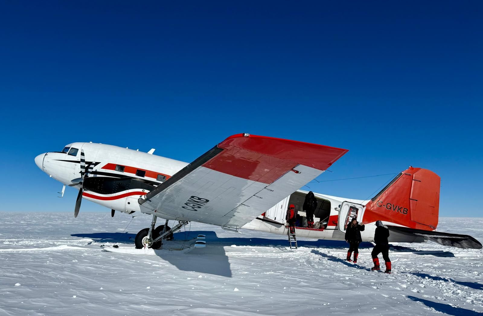 Red and white propeller plane on snowy ice with two people standing beside it under a clear blue sky