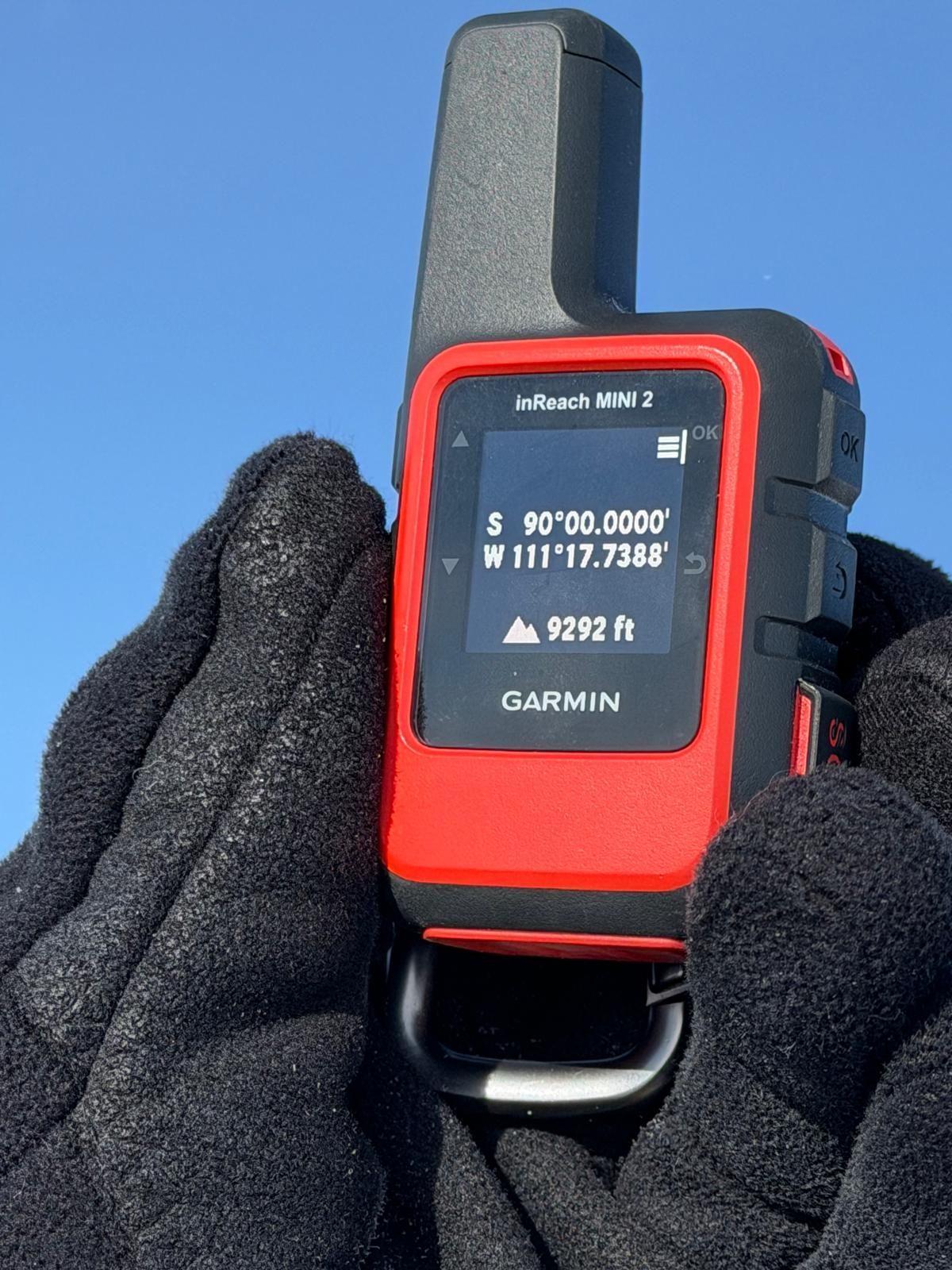 Gloved hand holding a red Garmin InReach Mini 2 displaying coordinates on its screen