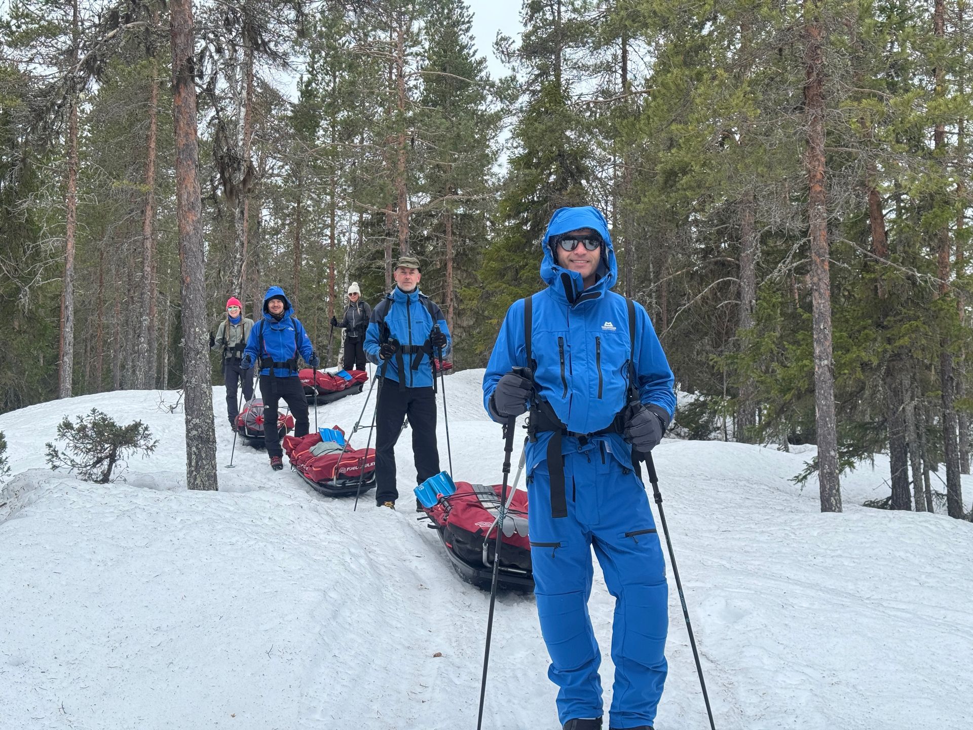 Three people in blue winter gear pulling sleds on a snowy forest trail