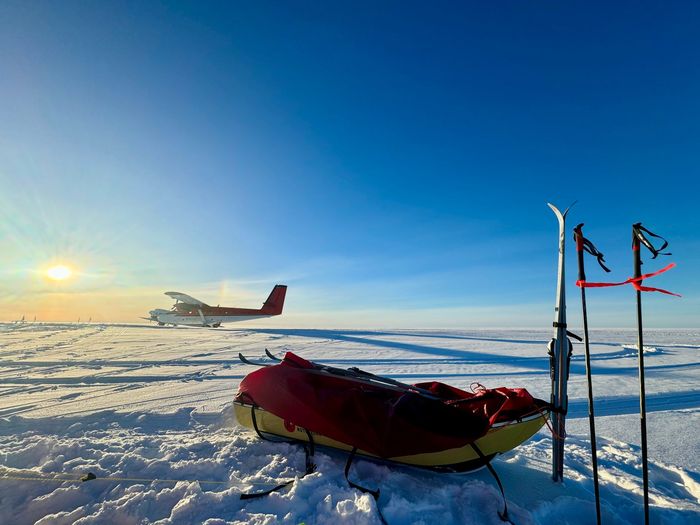 Red sled on snowy plain with skis and a small plane in the distance under a clear blue sky.