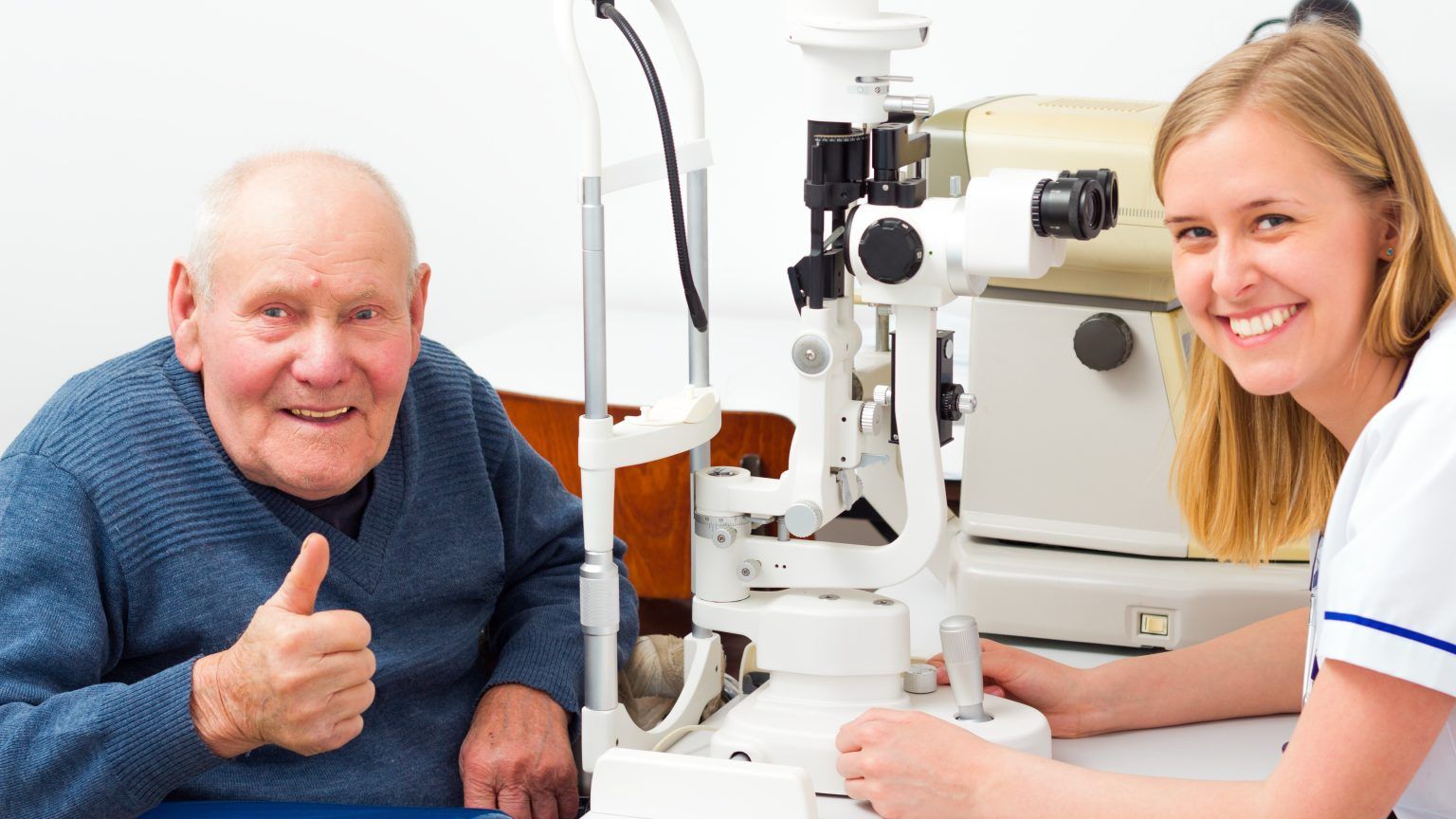 A smiling patient gives a thumbs-up while a professional prepares a slit lamp for an eye examination in a clinic.