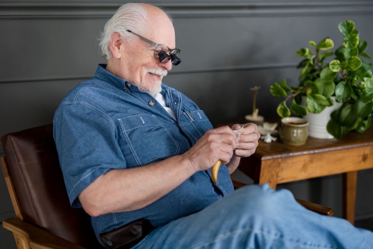 A person wearing specialized magnifying glasses works with a small, yellow object while sitting in a chair.