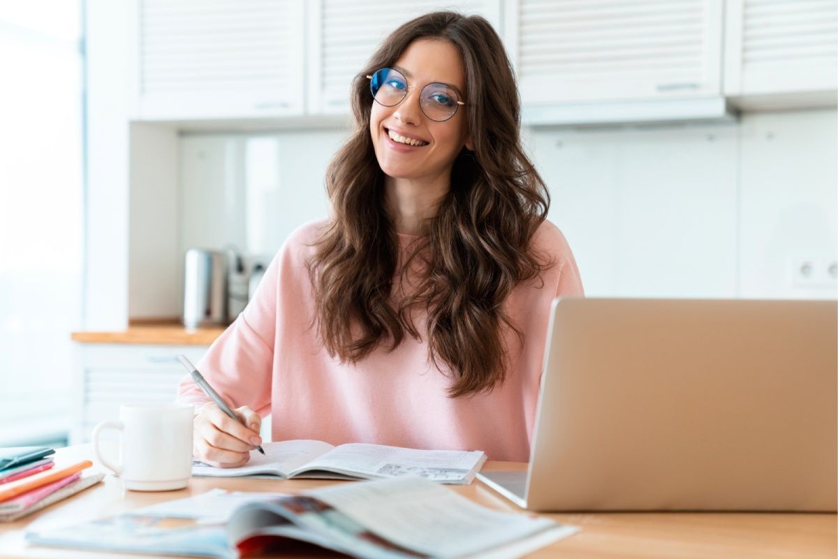 Woman in glasses smiles while working on a laptop, writing in a notebook in a kitchen.