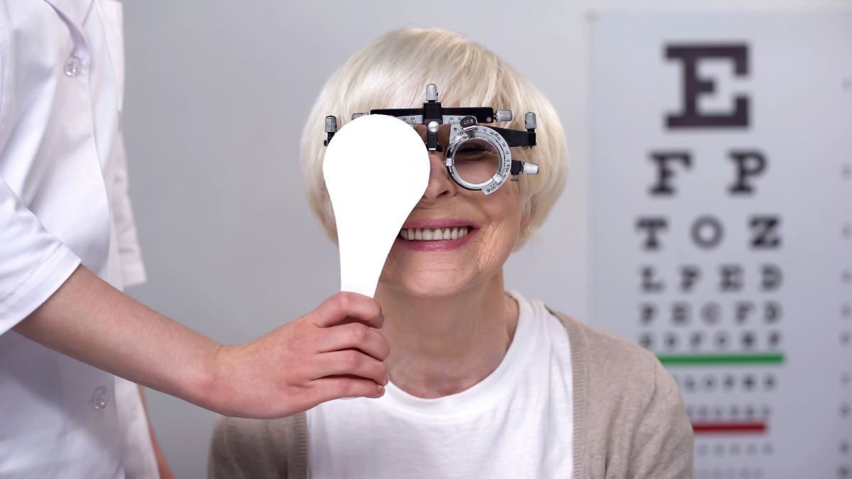 A healthcare provider holds an occluder over a patient's eye during an eye exam with a trial frame and vision chart.
