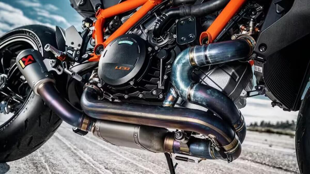 Primo piano del motore di una motocicletta con componenti arancioni, neri e in titanio su uno sfondo chiaro.