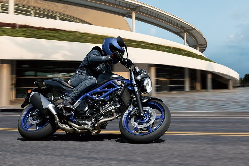 Motociclista su una Suzuki SV650 blu e nera su strada vicino a un edificio moderno.