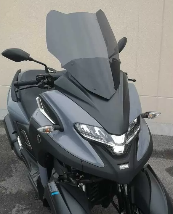 Scooter a tre ruote, grigio e nero, con parabrezza e specchietti oscurati, parcheggiato contro un muro beige.