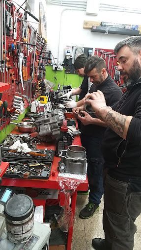 Tre meccanici lavorano su pezzi di motocicletta seduti al banco da lavoro di un'officina, con gli attrezzi appesi al muro.