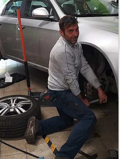 Un meccanico lavora su uno pneumatico per auto, in un'officina. È inginocchiato, usa una chiave inglese e sorride.