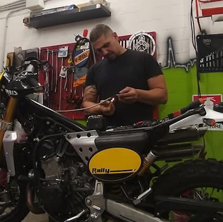Un uomo lavora su una motocicletta in un'officina. Indossa una camicia nera e tiene in mano un attrezzo.