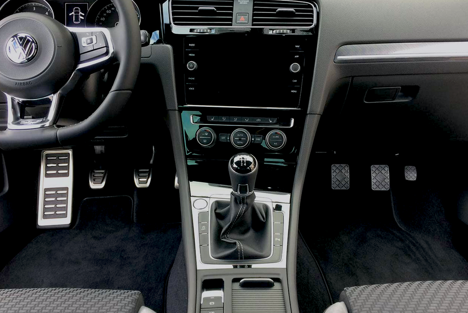 Vista interna del cruscotto di un'auto nera, con volante, schermo di infotainment, cambio e pedali.