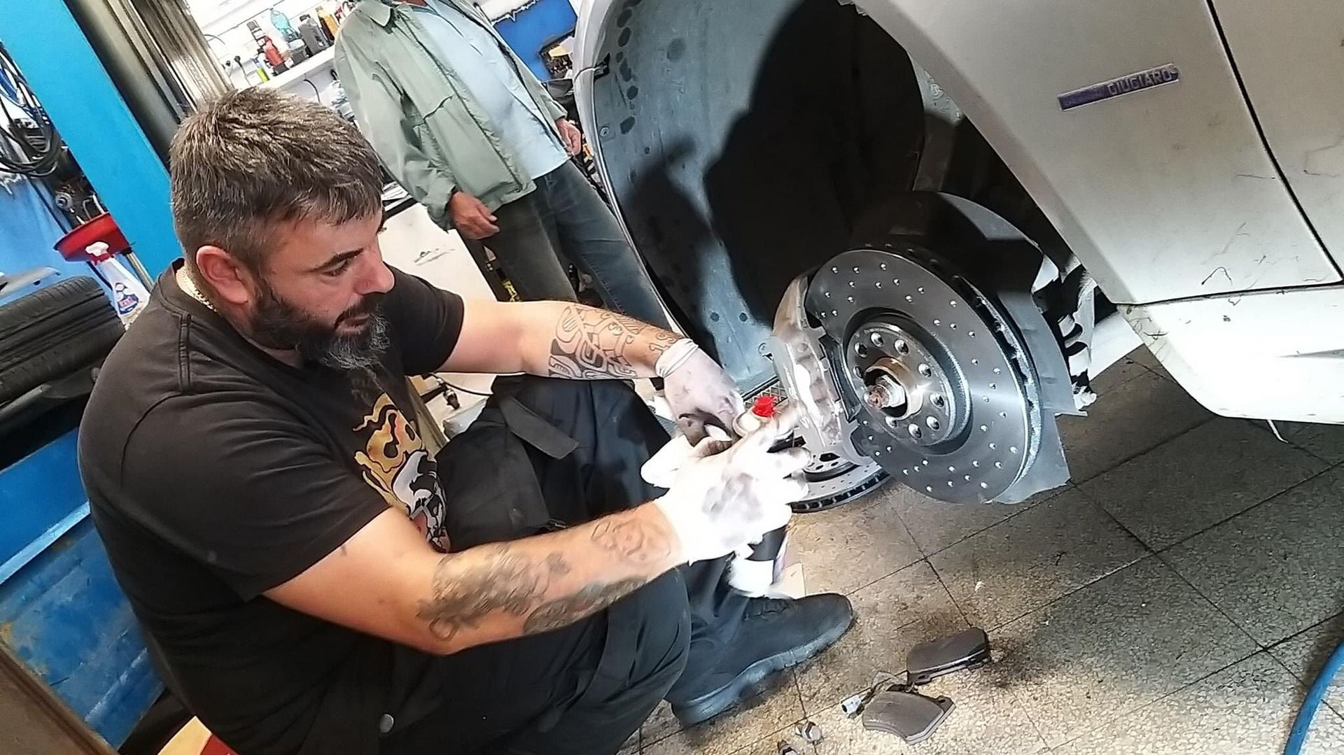 Un uomo con i guanti lavora sui freni di un'auto in un garage; un'altra persona è in piedi nelle vicinanze.
