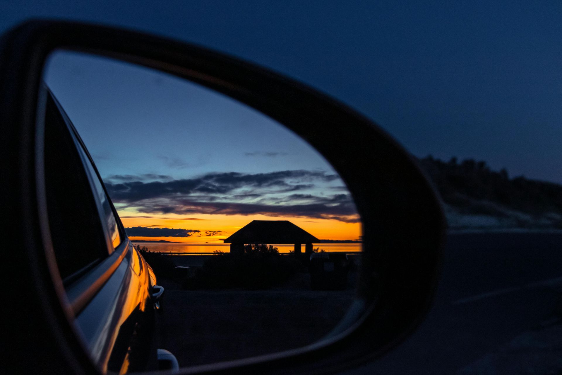 Il tramonto riflesso nello specchietto laterale di un'auto, con la sagoma di un edificio che si staglia contro il cielo.