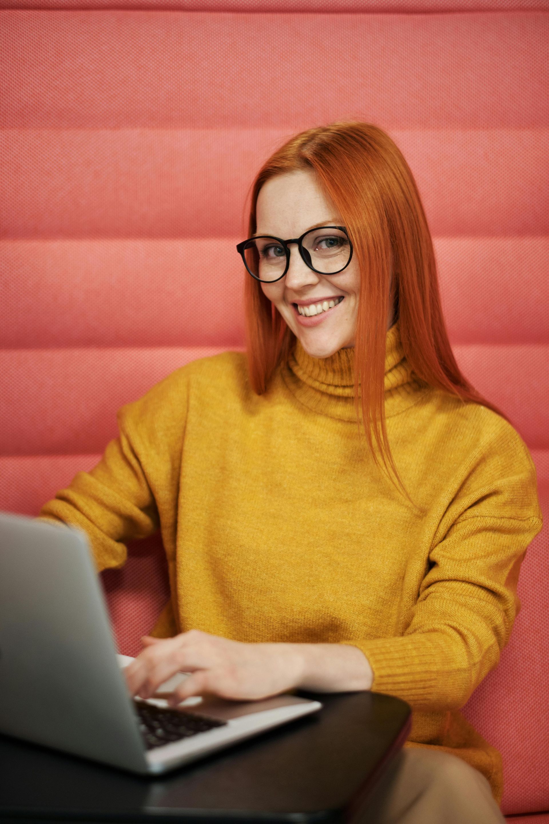 Donna con i capelli rossi e gli occhiali che usa un computer portatile, sorride alla telecamera, indossa un maglione gialla.