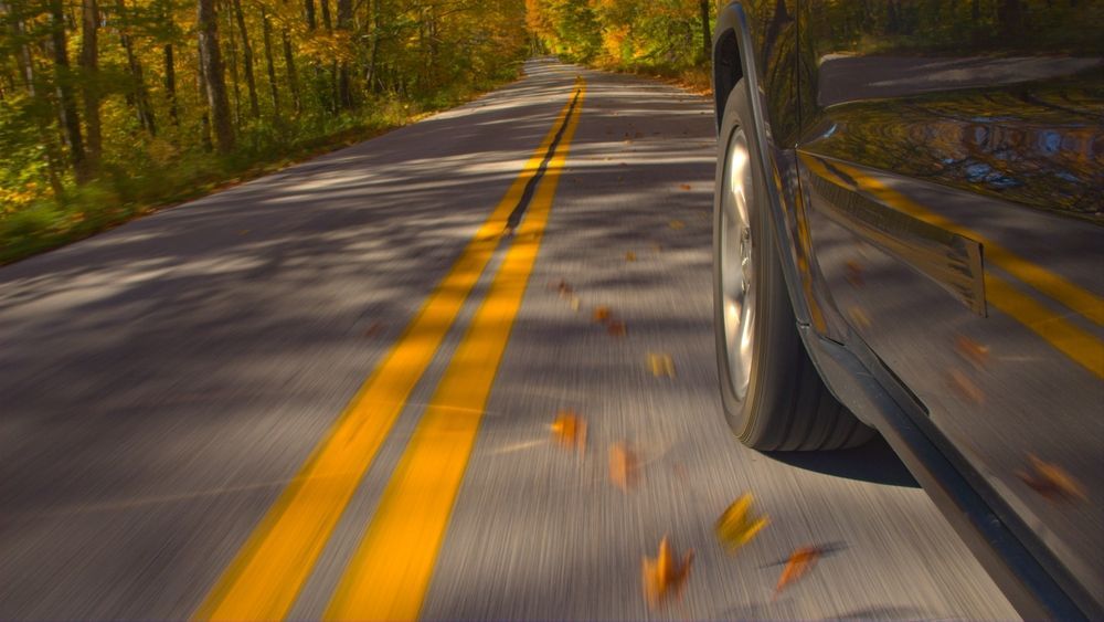 A car is driving down a road with leaves falling on the ground.