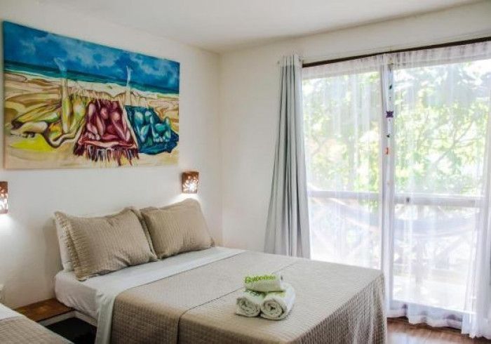Quarto com cama de casal, obras de arte com temática de praia e porta de vidro deslizante com cortinas translúcidas.