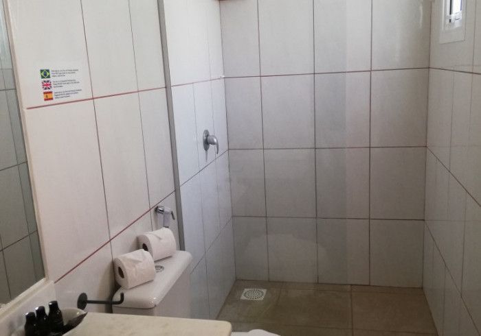 Um pequeno box de chuveiro revestido com azulejos brancos em um banheiro, com um chuveiro fixo e dois rolos de papel higiênico.