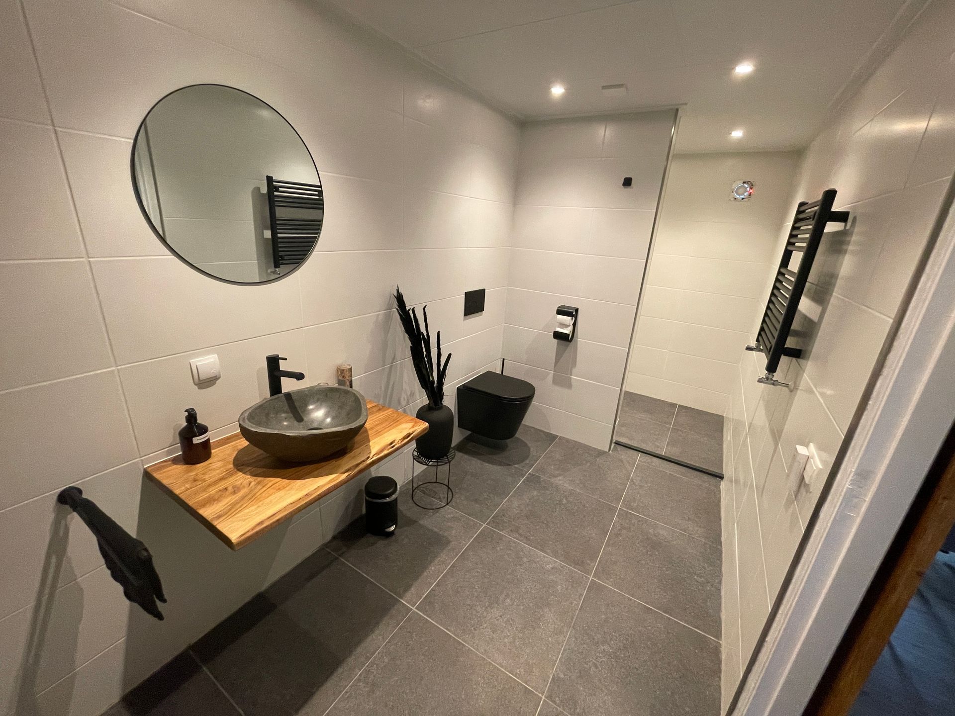 Moderne badkamer met grijze tegels, zwarte kranen, houten aanrechtblad en ronde spiegel.