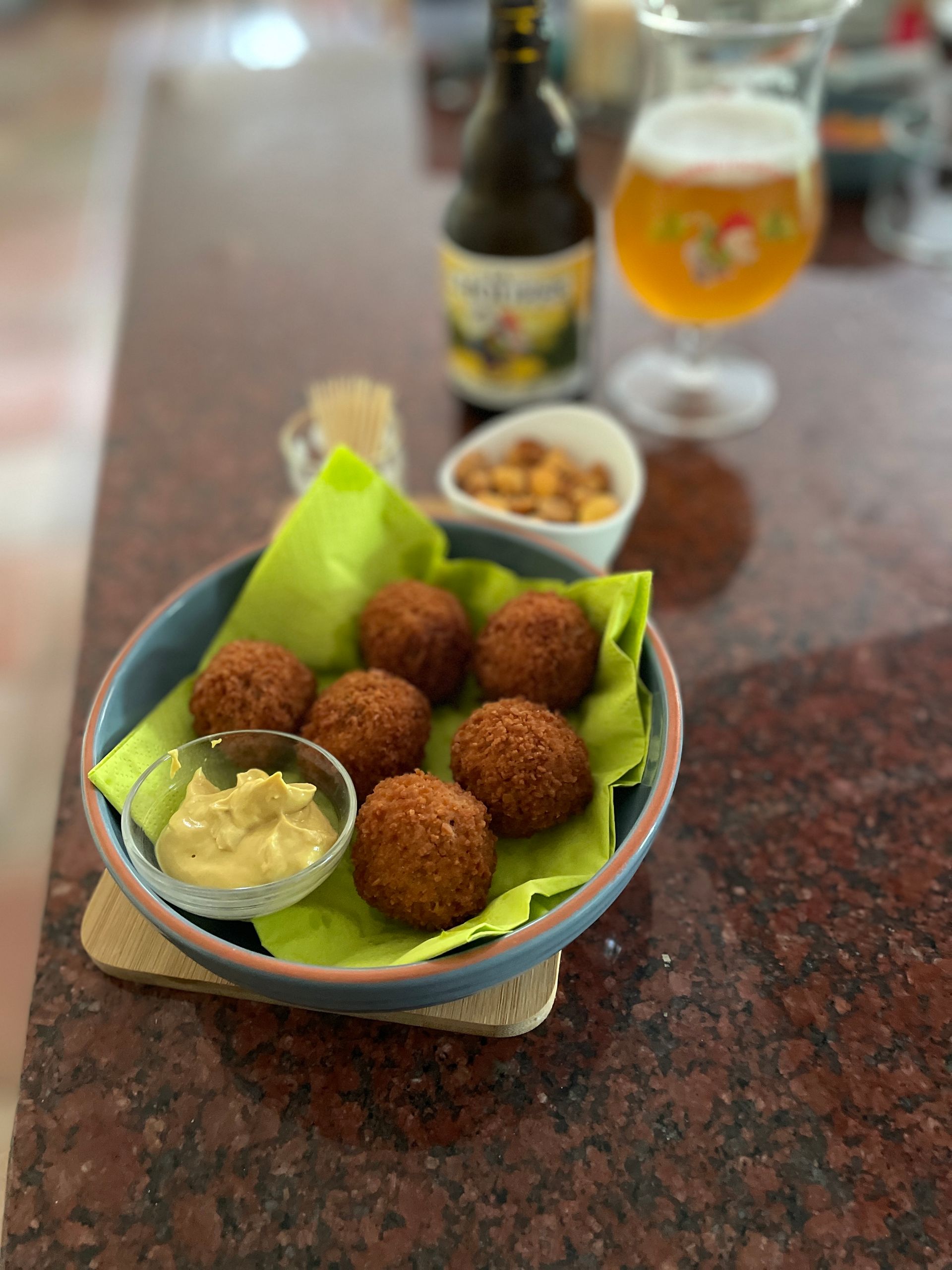 Een bord met gefrituurde balletjes, mosterd, noten, een flesje bier en een glas bier op de toonbank.