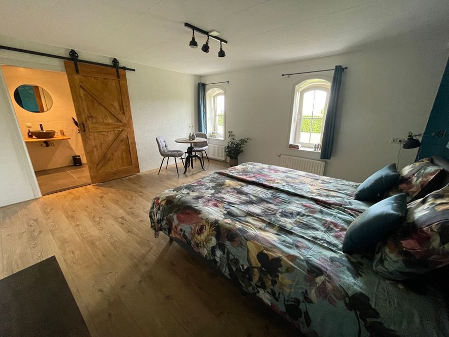 Slaapkamer met houten vloer, bed met bloemensprei, schuifdeur naar de badkamer, klein tafeltje en stoelen.