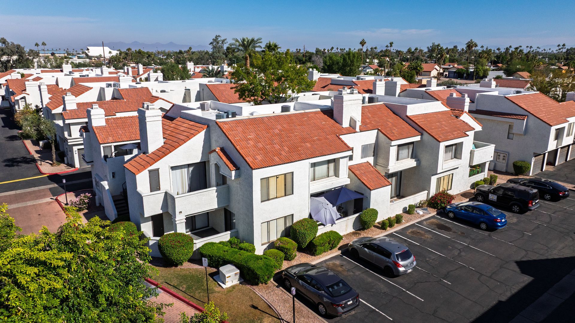 Apartment complex with white stucco walls, orange tile roofs, palm trees, and parked cars. Talavera Apartments features refined apartments in Tempe AZ—explore today.