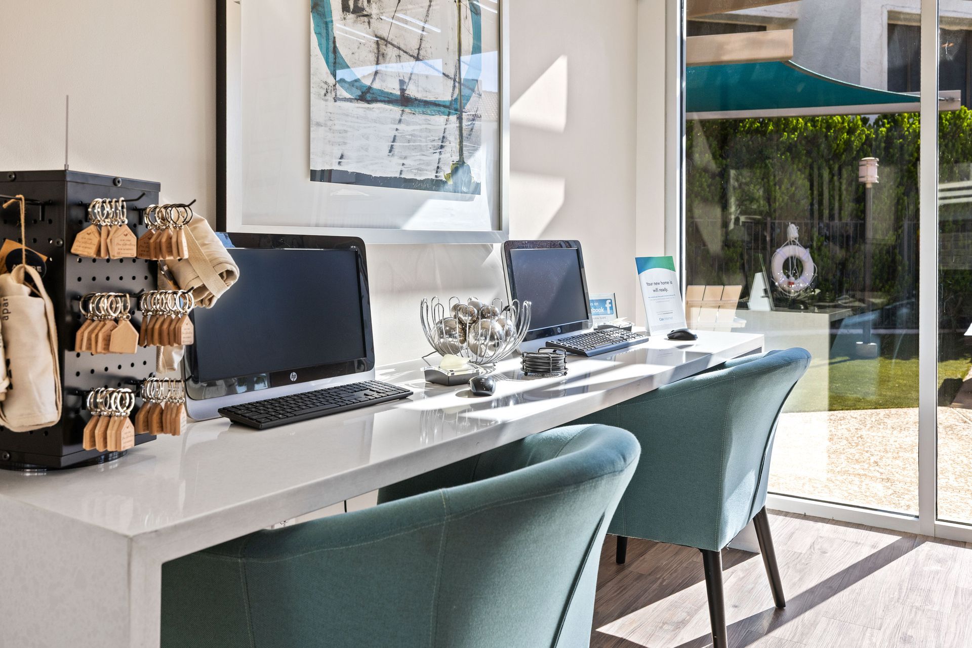 Two teal chairs at a white desk with two computers, key rack, and a view of an outdoor area. Discover convenient Tempe AZ apartment rentals at Talavera Apartments today.