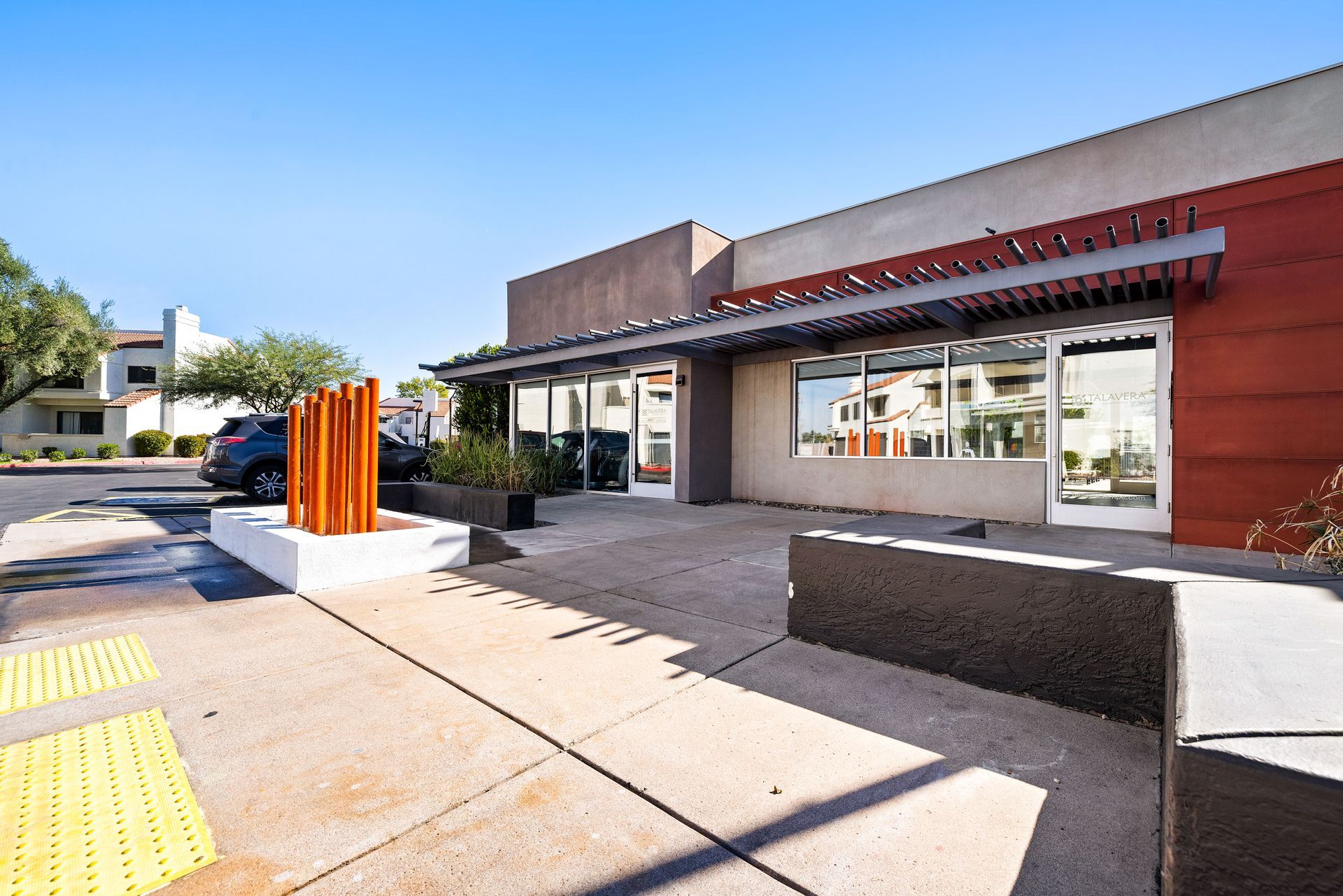 Modern building with large windows and a shaded entryway. Sidewalk with yellow tactile paving. Explore stylish Tempe, Arizona apartments at Talavera Apartments today.