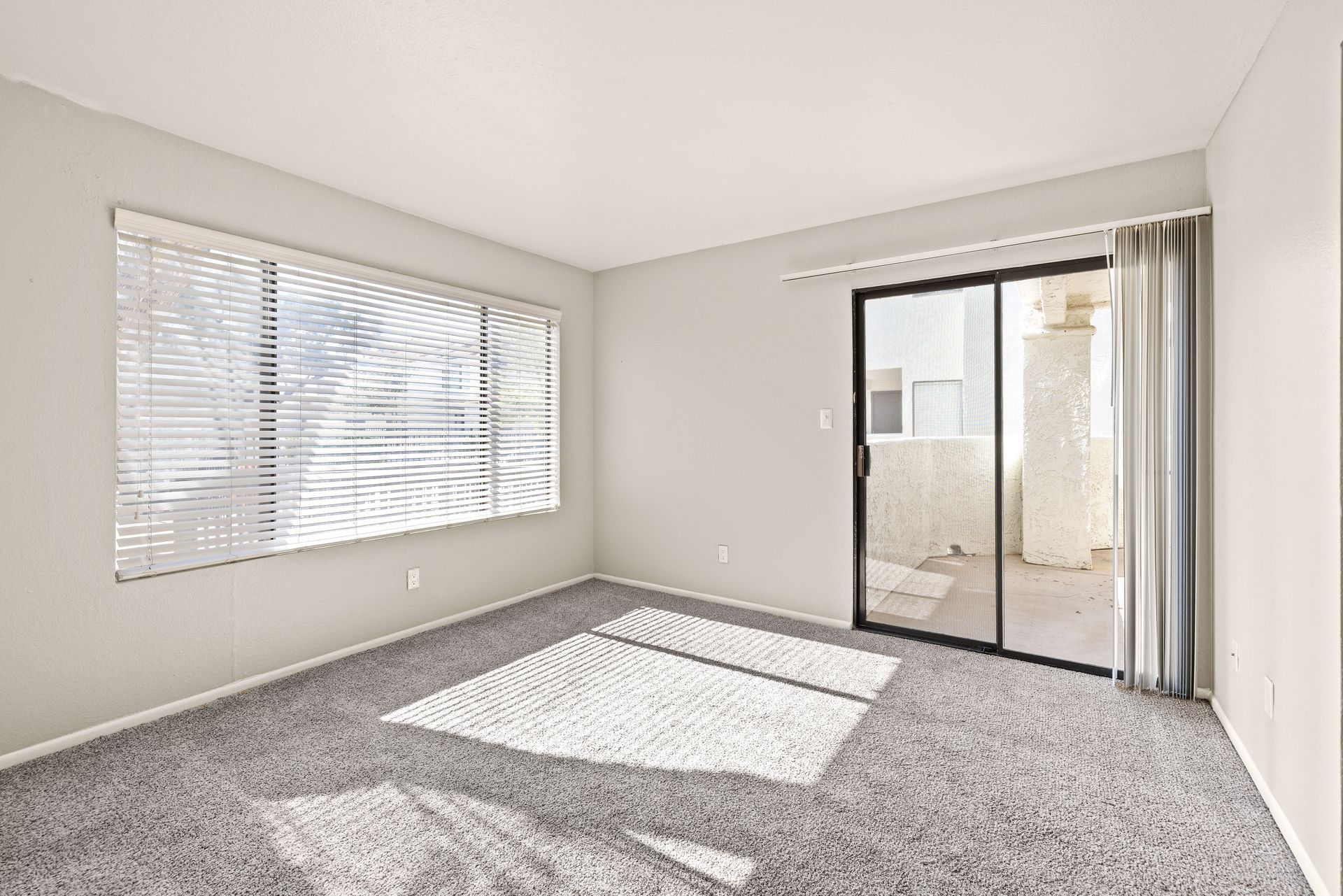 Empty room with gray carpet, window with blinds, and sliding glass door to a balcony. Talavera Apartments places you close to it all with downtown Tempe apartments—learn more.