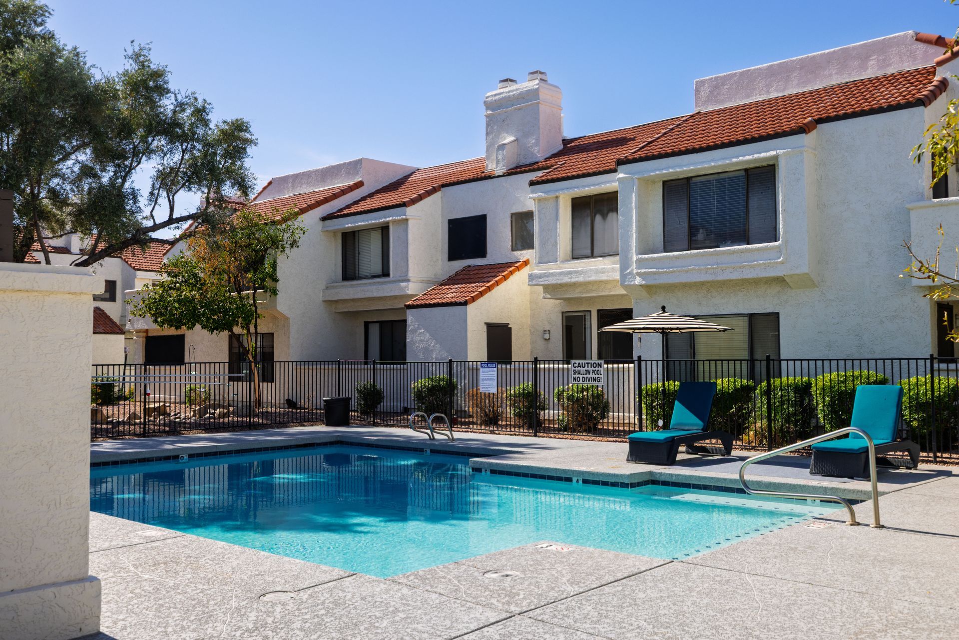 Swimming pool in front of white stucco apartment building with red tile roof; blue lounge chairs. Talavera Apartments provides welcoming apartment homes in Tempe AZ—see more.