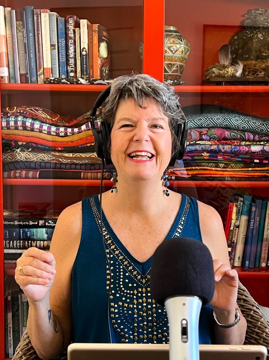 Woman with gray hair wearing headphones and turquoise top, smiling in front of a bookshelf and microphone.