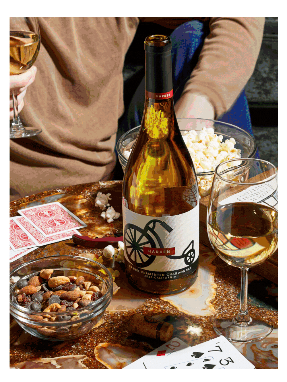 Wine bottle with bicycle label, glasses of wine, snacks, and playing cards on a table.