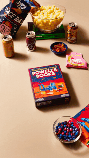 Snacks and drinks surrounding a book; chips, popcorn, candy, and soda cans on a light surface.