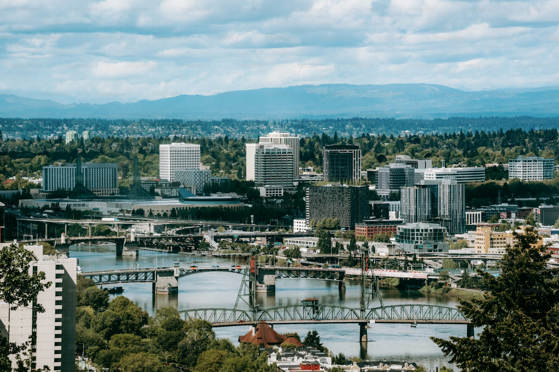 City skyline with bridges over a river, trees in foreground, mountains in the distance under cloudy sky.