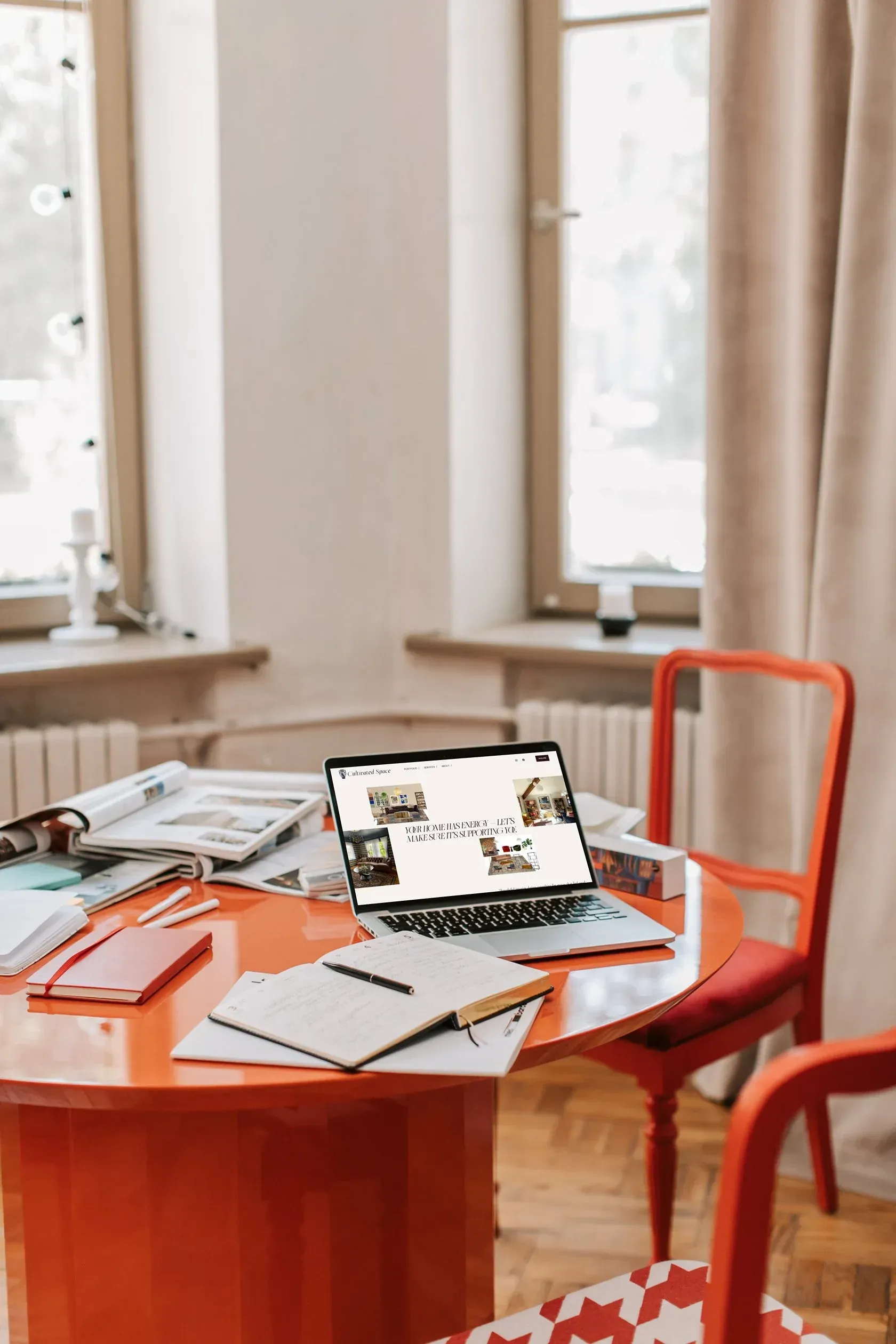 Red chair by a round orange table with an open laptop in a bright room with large windows