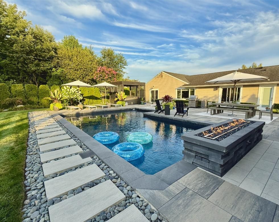 Backyard pool with stone patio, fire pit, lounge chairs, and blue pool floats under a sunny sky.