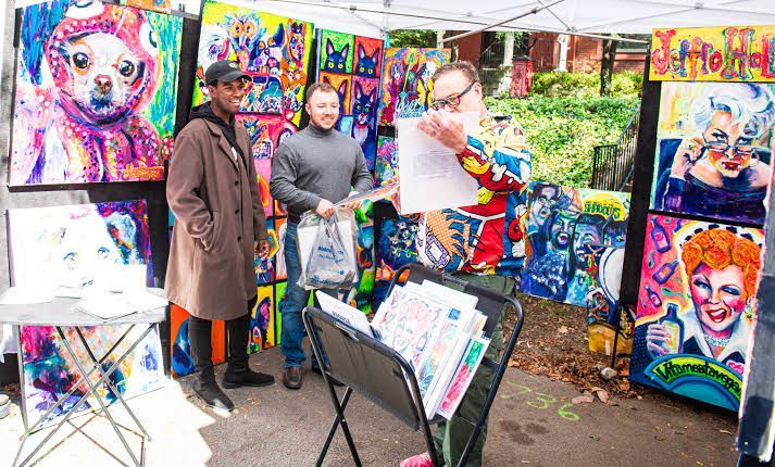Outdoor art booth with colorful portrait paintings displayed and two people browsing under a canopy
