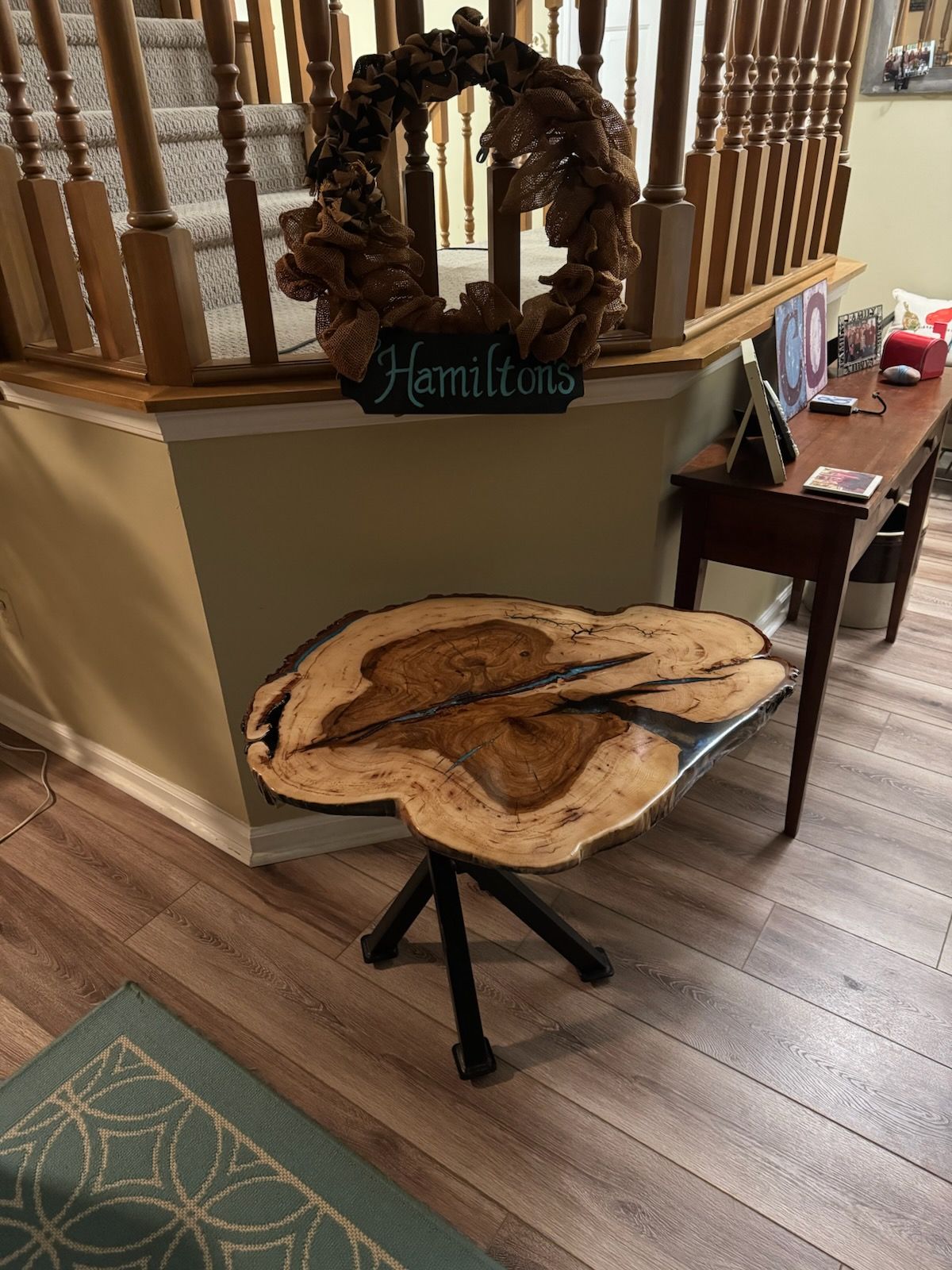 Decorative wood slice table beside a staircase and railing, with a wreath and “Hamilton” sign nearby