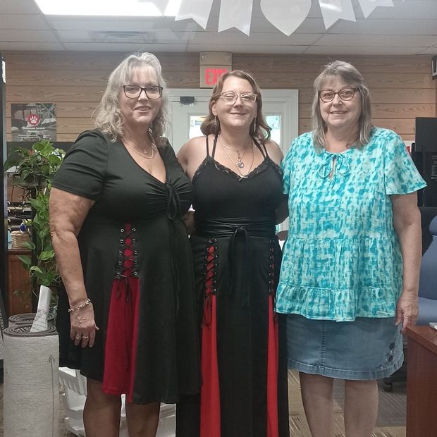 Three people standing indoors, smiling, in black, red, and teal outfits.