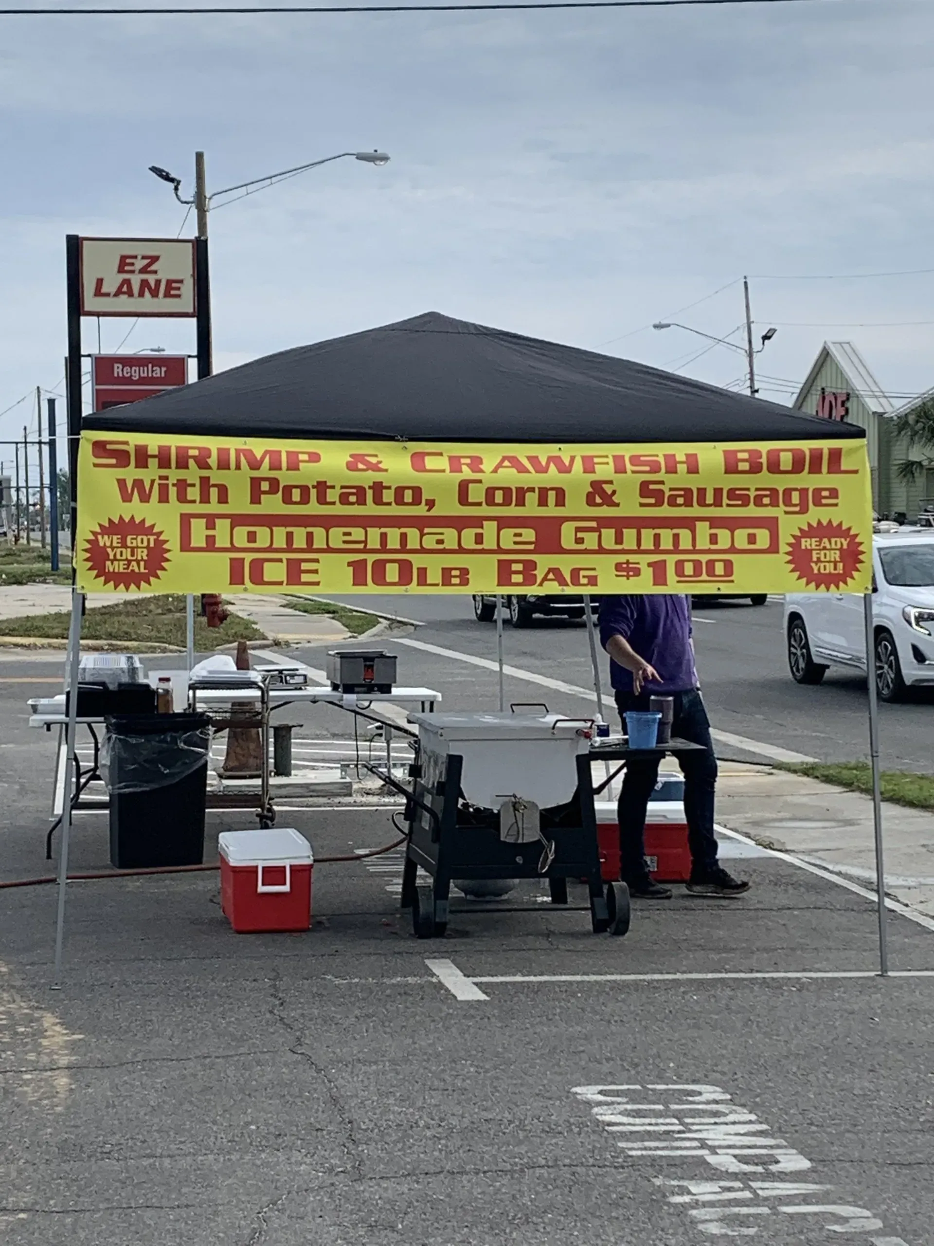 A man stands under a tent with a sign that says shrimp and crawfish boil with potato corn and sausage
