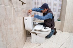 Plumber repairing a toilet in a tiled bathroom, adjusting the tank with tools.
