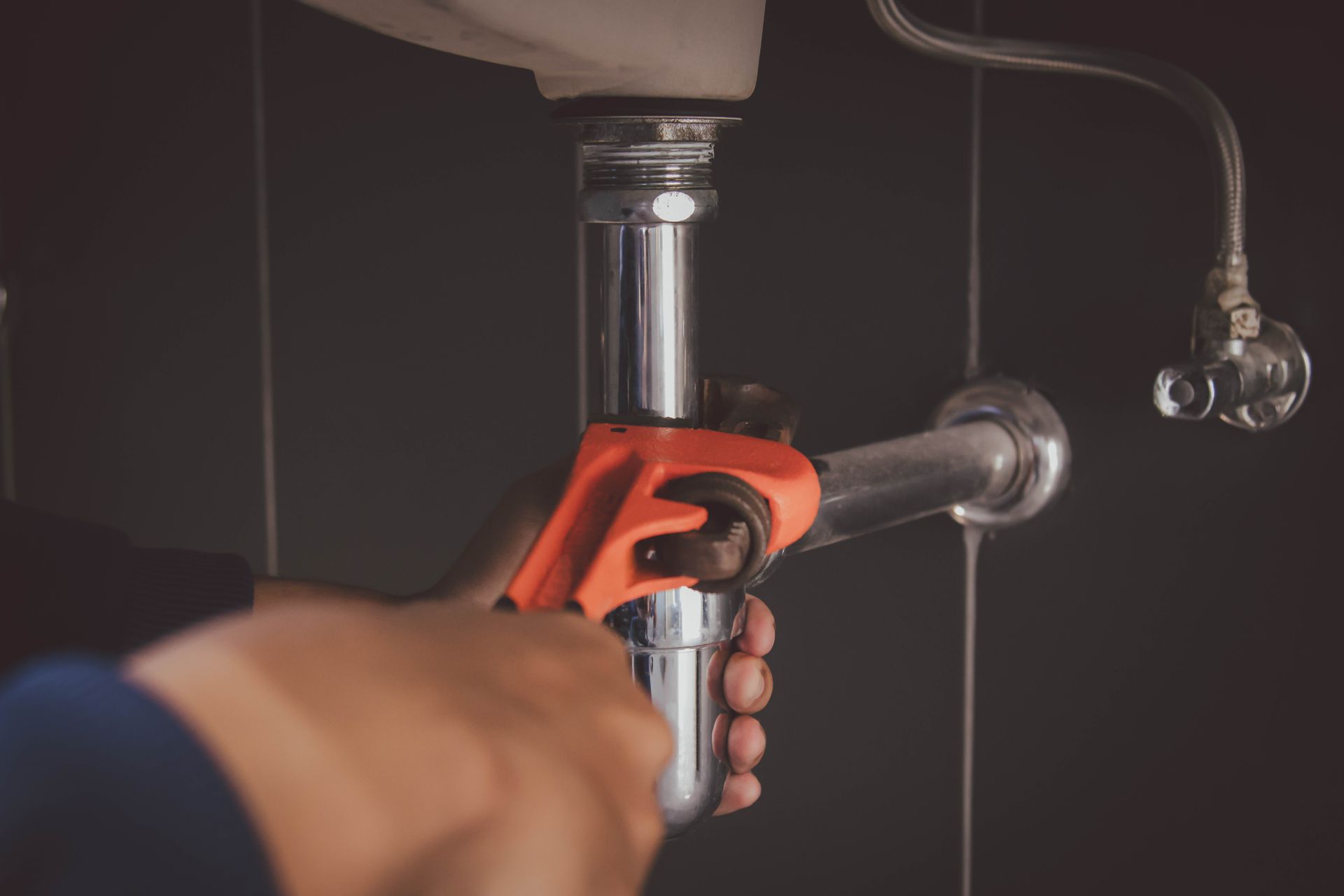 Hands tightening a red pipe wrench on a chrome sink drain pipe under a sink.