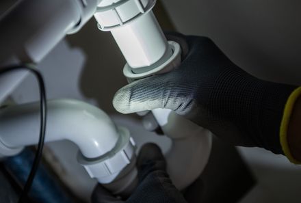 Gloved hands tightening a white PVC pipe under a sink with a wrench